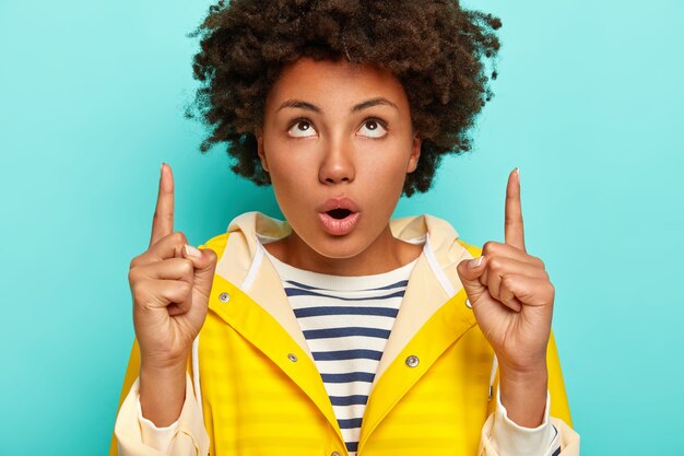 Mujer joven sorprendida con puntos de corte de pelo Afro arriba, abre la boca, enfocada hacia arriba con sorpresa, vestida con suéter a rayas e impermeable amarillo, aislado sobre fondo azul.