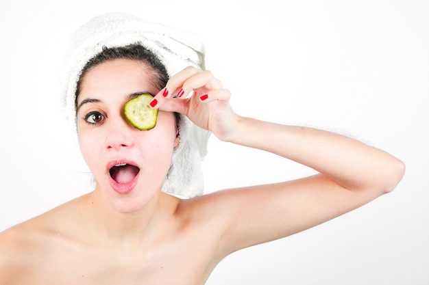 Mujer joven sorprendida que lleva a cabo la rebanada del pepino sobre sus ojos aislados en el fondo blanco