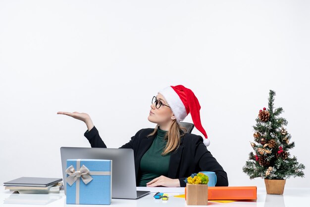 Mujer joven sorprendida con sombrero de santa claus y anteojos sentado en una mesa con un árbol de Navidad y un regalo y apuntando hacia el lado derecho sobre fondo blanco.