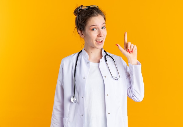 Mujer joven sorprendida en uniforme médico con estetoscopio apuntando hacia arriba