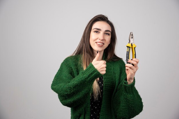 Mujer joven sosteniendo alicates y mostrando un pulgar hacia arriba.