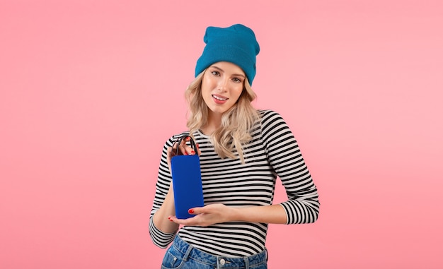 Mujer joven sosteniendo altavoz inalámbrico escuchando música vistiendo camisa a rayas y sombrero azul sonriendo posando en rosa