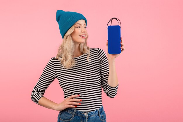 Mujer joven sosteniendo altavoz inalámbrico escuchando música vistiendo camisa a rayas y sombrero azul sonriendo posando en rosa