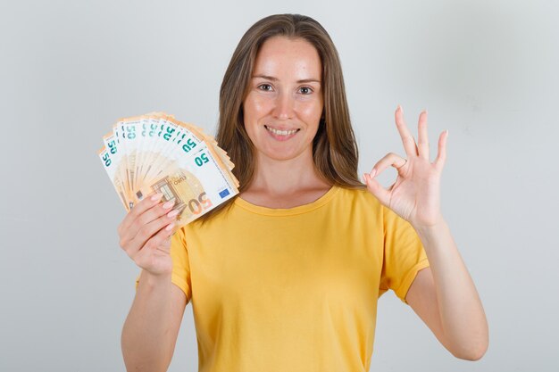 Mujer joven sosteniendo billetes en euros con signo ok en camiseta amarilla y mirando feliz