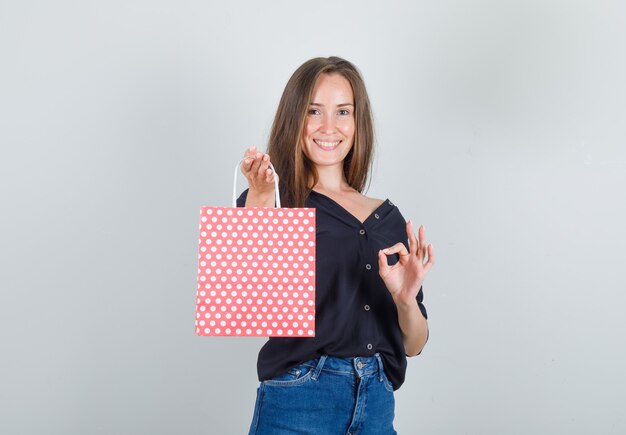 Mujer joven sosteniendo una bolsa de papel con signo ok en camisa negra, pantalones cortos de jeans y mirando alegre