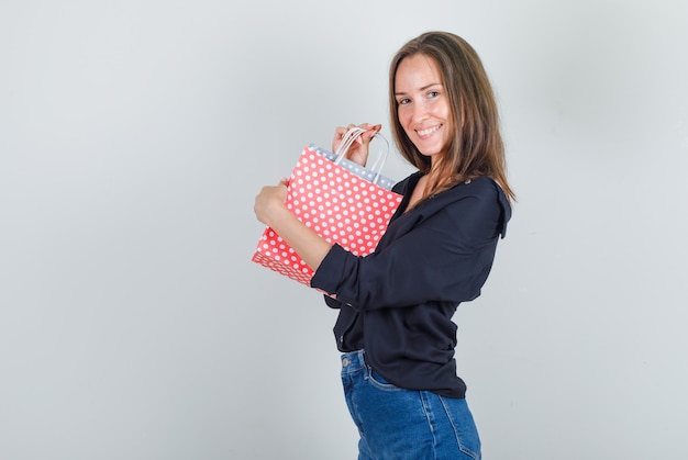 Mujer joven sosteniendo bolsas de papel en camisa negra, pantalones cortos de jeans y mirando alegre.