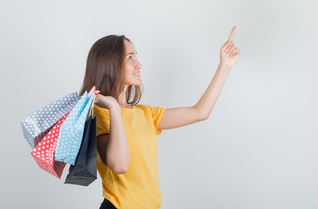 Mujer joven sosteniendo bolsas de papel con el dedo hacia arriba en camiseta amarilla, pantalones y mirando contento.