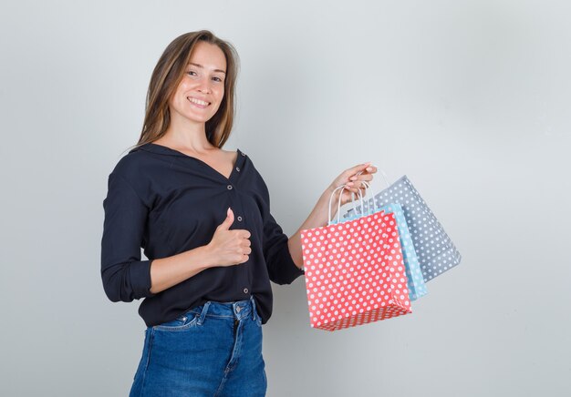 Mujer joven sosteniendo bolsas de papel con el pulgar hacia arriba en camisa negra, pantalones vaqueros y mirando alegre