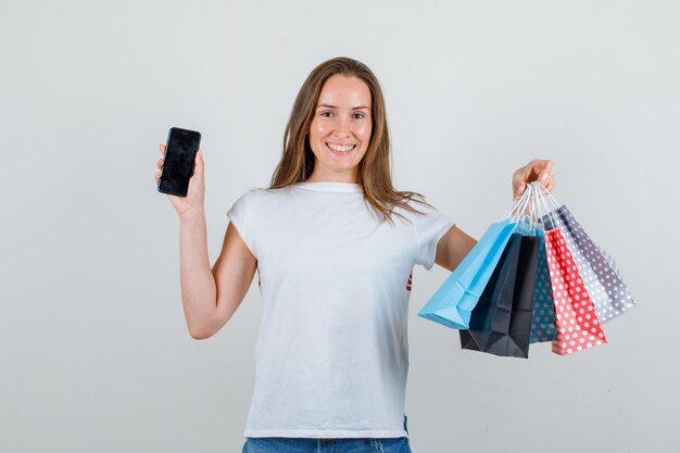 Mujer joven sosteniendo bolsas de papel y smartphone en camiseta blanca, pantalones cortos y mirando alegre