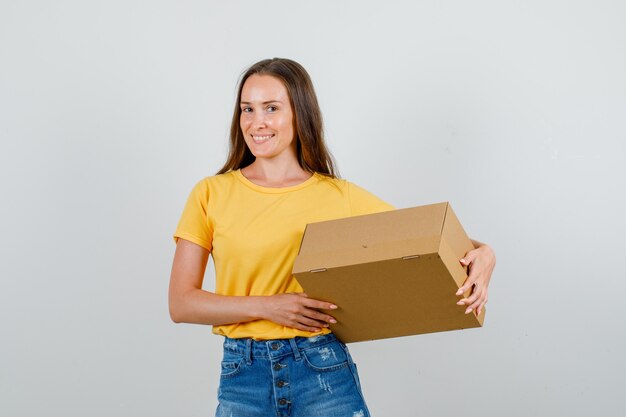 Mujer joven sosteniendo una caja de cartón y sonriendo en camiseta, vista frontal de pantalones cortos.