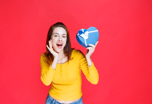 Mujer joven sosteniendo una caja de regalo con forma de corazón azul y sintiéndose sorprendido