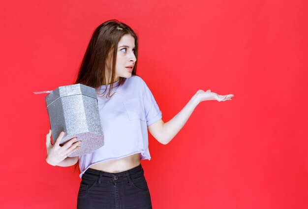 mujer joven sosteniendo una caja de regalo plateada y apuntando a alguien.