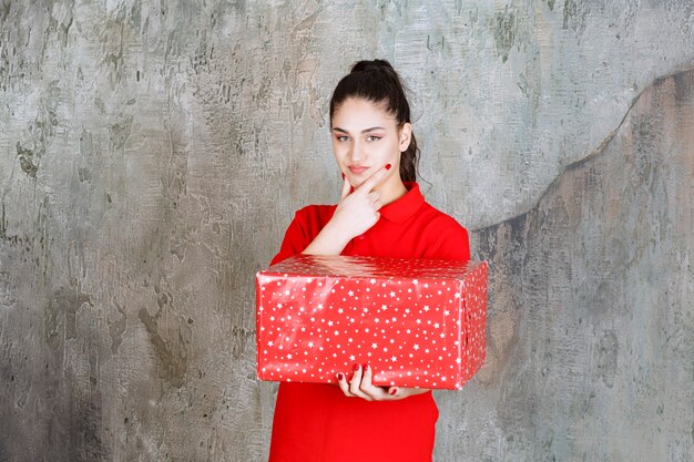 Mujer joven sosteniendo una caja de regalo roja con puntos blancos y se ve pensativa.