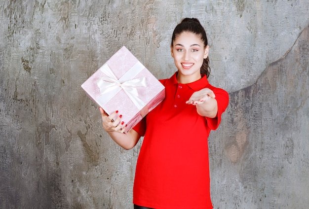 Mujer joven sosteniendo una caja de regalo rosa envuelta con una cinta blanca y llamando a la persona que está adelante para presentarla.