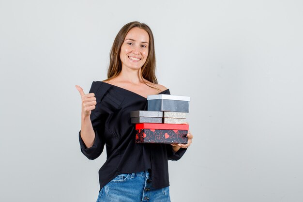 Mujer joven sosteniendo cajas presentes con el pulgar hacia arriba en camisa, pantalones cortos y mirando feliz. vista frontal.