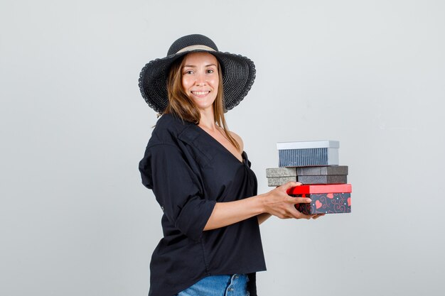 Mujer joven sosteniendo cajas de regalo en camisa, pantalones cortos, sombrero y mirando alegre