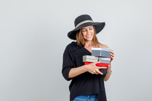 Mujer joven sosteniendo cajas de regalo en camisa, pantalones cortos, sombrero y mirando alegre