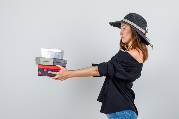 Mujer joven sosteniendo cajas de regalo en camisa, pantalones cortos, sombrero y mirando serio.