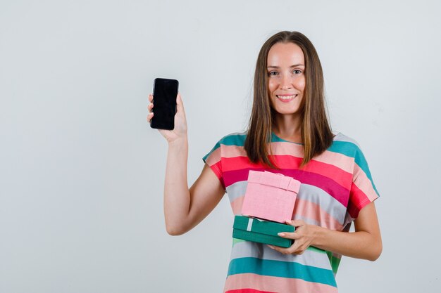 Mujer joven sosteniendo cajas de regalo y teléfono móvil en camiseta y mirando contento, vista frontal.