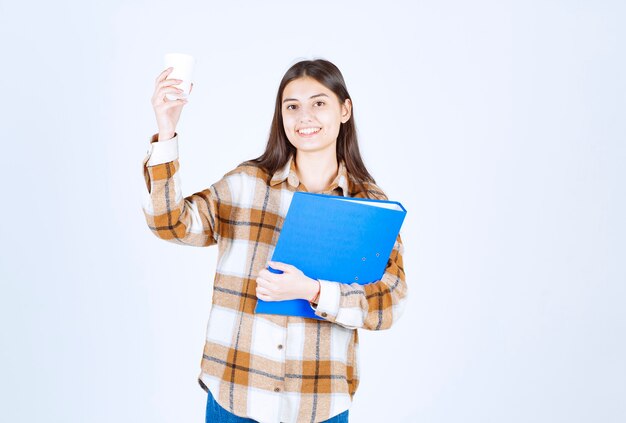 Mujer joven sosteniendo una carpeta azul y una taza de café en la pared blanca.