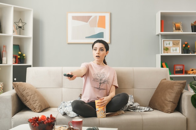 Mujer joven sosteniendo el control remoto del televisor a la cámara sentado en el sofá detrás de la mesa de café en la sala de estar