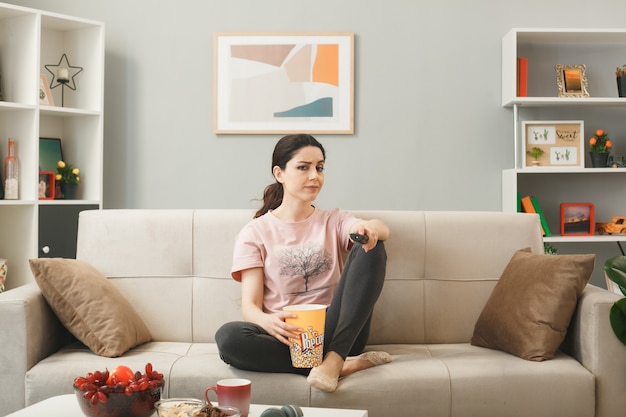 Mujer joven sosteniendo el control remoto del televisor sentado en el sofá detrás de la mesa de café en la sala de estar