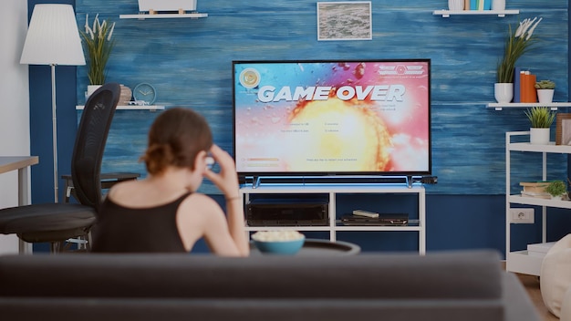 Foto gratuita mujer joven sosteniendo un controlador inalámbrico jugando al tirador espacial de acción decepcionada porque perdió el juego en línea en la consola frente a la televisión. chica jugadora sentada en el sofá descontenta con el rendimiento del juego.