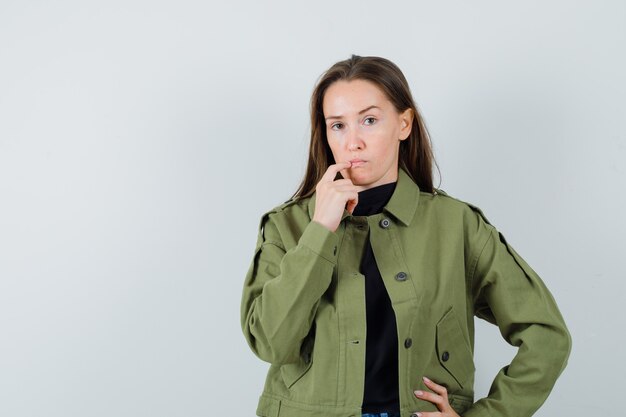 Mujer joven sosteniendo el dedo en la boca con chaqueta verde y mirando confundido, vista frontal.