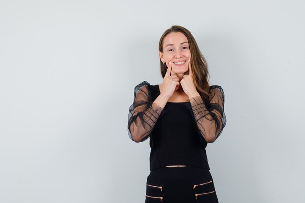 Mujer joven sosteniendo los dedos índices cerca de la boca, forzando una sonrisa en blusa negra y pantalón negro y luciendo feliz, vista frontal.