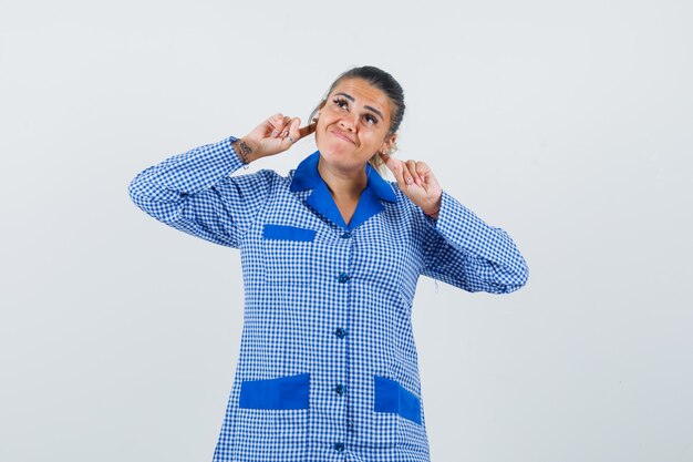 Mujer joven sosteniendo los dedos índices detrás de la oreja en camisa de pijama a cuadros azul y mirando bonita vista frontal.