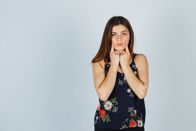 Mujer joven sosteniendo los dedos en las mejillas en blusa y mirando triste, vista frontal.