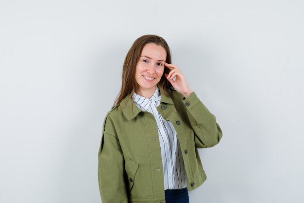 Mujer joven sosteniendo los dedos en las sienes en camisa, chaqueta y mirando confiado. vista frontal.