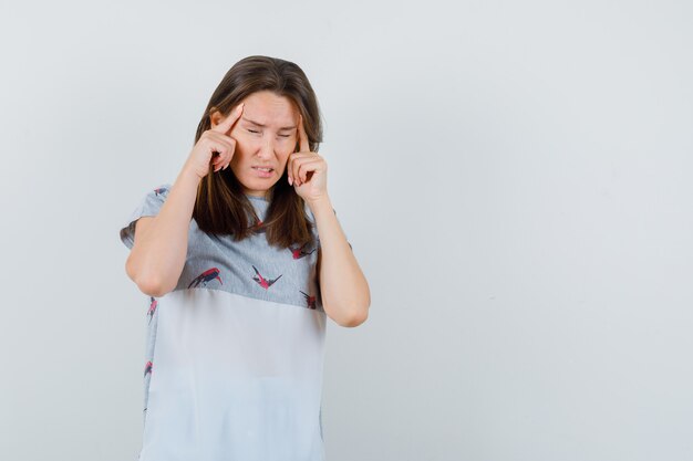 Mujer joven sosteniendo los dedos en las sienes y tener dolor de cabeza en la vista frontal de la camiseta.