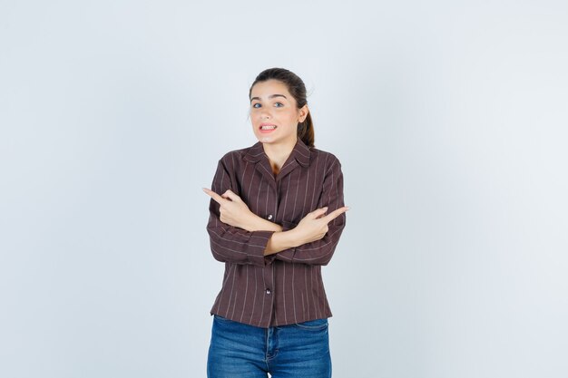 Mujer joven sosteniendo dos brazos cruzados, apuntando en direcciones opuestas con los dedos índices en camisa a rayas, jeans y mirando feliz, vista frontal.