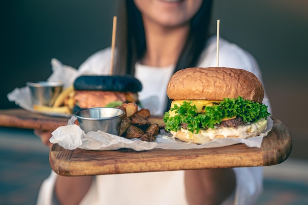 Foto gratuita una mujer joven sosteniendo hamburguesas de comida rápida en una bandeja de madera al aire libre