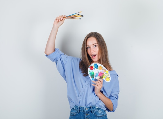 Mujer joven sosteniendo herramientas de pintura en camisa azul, pantalones cortos y mirando feliz.