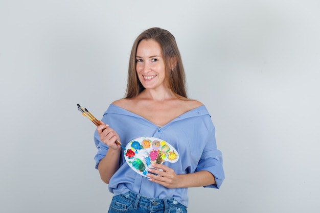 Mujer joven sosteniendo herramientas de pintura en camisa, pantalones cortos y mirando alegre