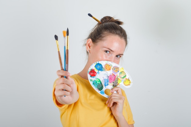 Mujer joven sosteniendo herramientas de pintura en camiseta amarilla y mirando feliz