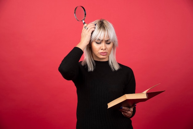 Mujer joven sosteniendo un libro con lupa en una pared roja.