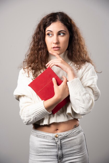 Mujer joven sosteniendo un libro rojo sobre una pared gris. Foto de alta calidad