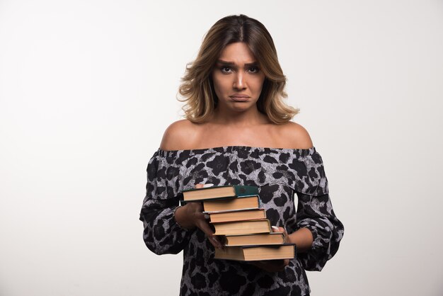 Mujer joven sosteniendo libros mientras está triste en la pared blanca.