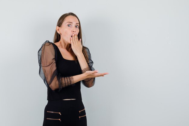 Mujer joven sosteniendo la mano en la boca mientras muestra algo a la izquierda en blusa negra y mirando sorprendido