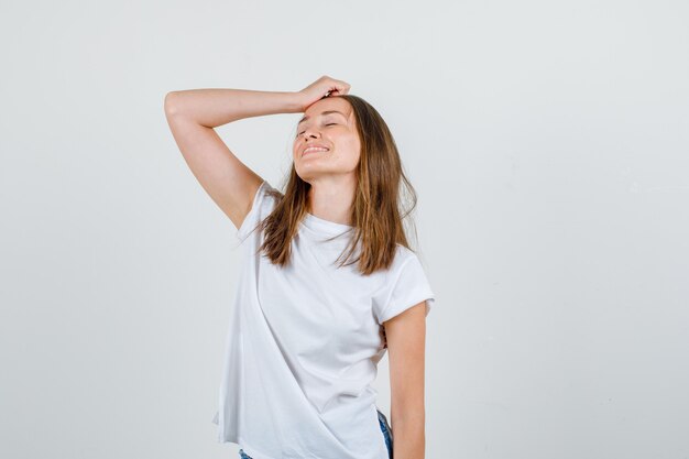 Mujer joven sosteniendo la mano en la cabeza en camiseta blanca, pantalones cortos y mirando esperanzado