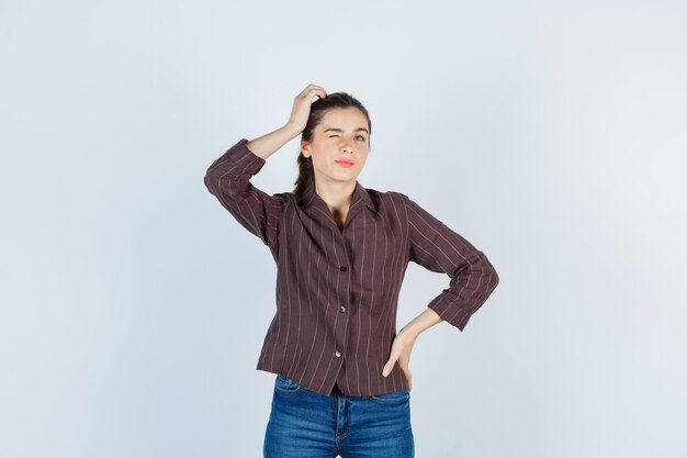 Mujer joven sosteniendo una mano en la cabeza, poniendo la mano en la cadera, pensando en algo en camisa a rayas, jeans y mirando pensativo. vista frontal.