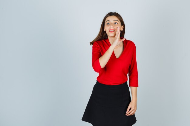 Mujer joven sosteniendo la mano cerca de la boca como contando secreto en blusa roja