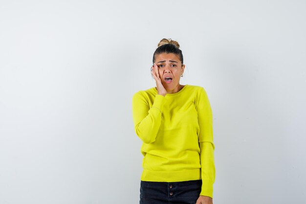 Mujer joven sosteniendo la mano cerca de la boca, manteniendo la boca abierta en suéter amarillo y pantalón negro y mirando sorprendido