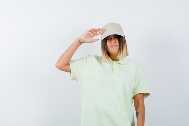 Mujer joven sosteniendo la mano cerca de la cabeza para saludar con camiseta, gorra y parecer confiado. vista frontal.