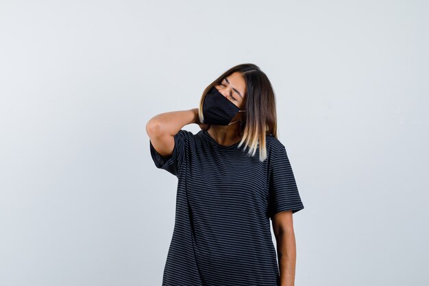 Mujer joven sosteniendo la mano en el cuello, con dolor de cuello en vestido negro, máscara negra y mirando exhausto, vista frontal.