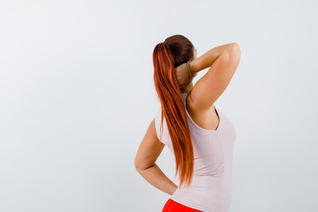 Mujer joven sosteniendo la mano en el cuello mientras mantiene la mano en la cintura en camiseta y parece segura, vista posterior.