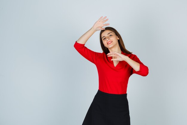 Mujer joven sosteniendo la mano debajo de la barbilla, agitando la mano para saludar en blusa roja